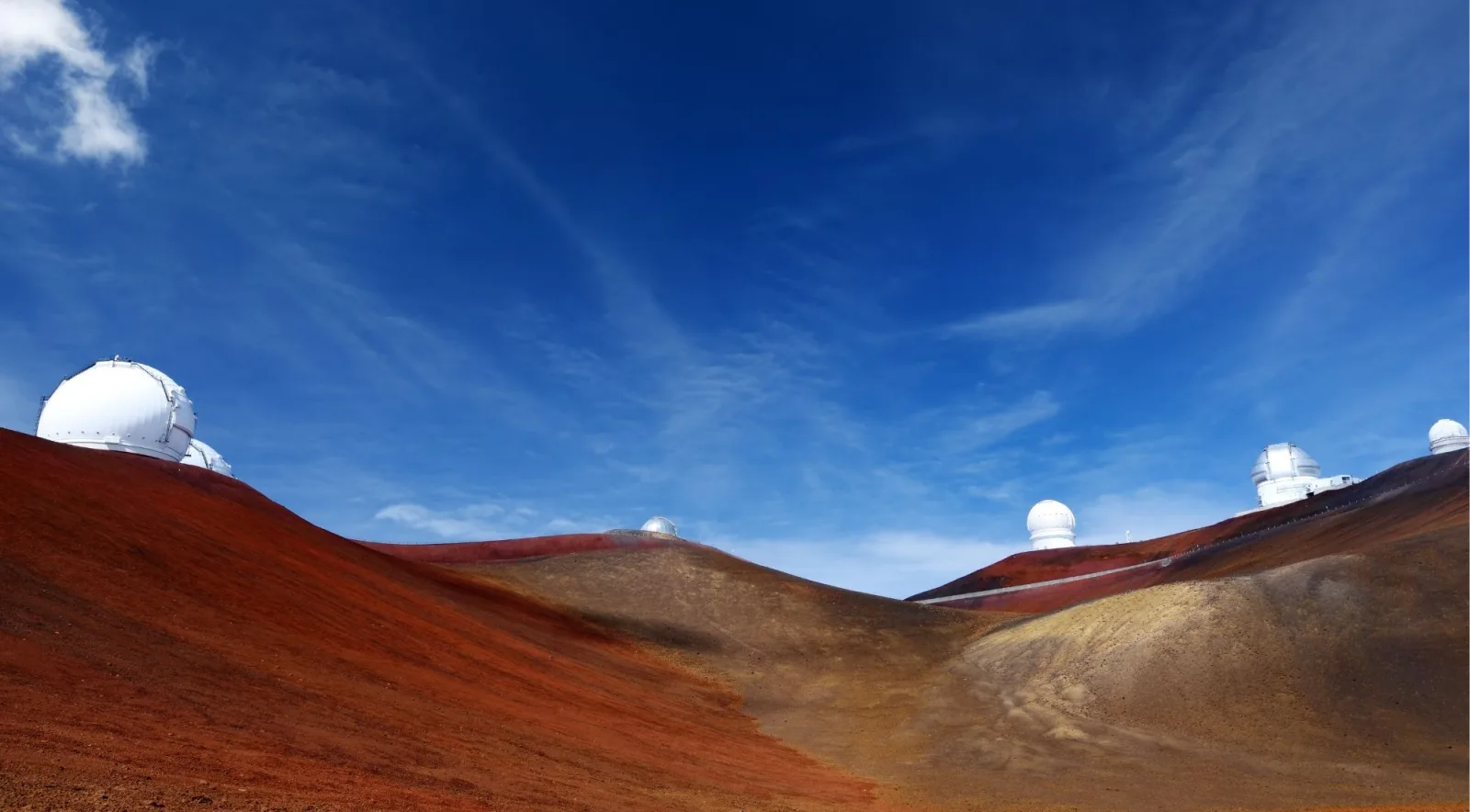 Panoramic view of Mauna Kea's red cinder summit with multiple white observatory domes along the ridge