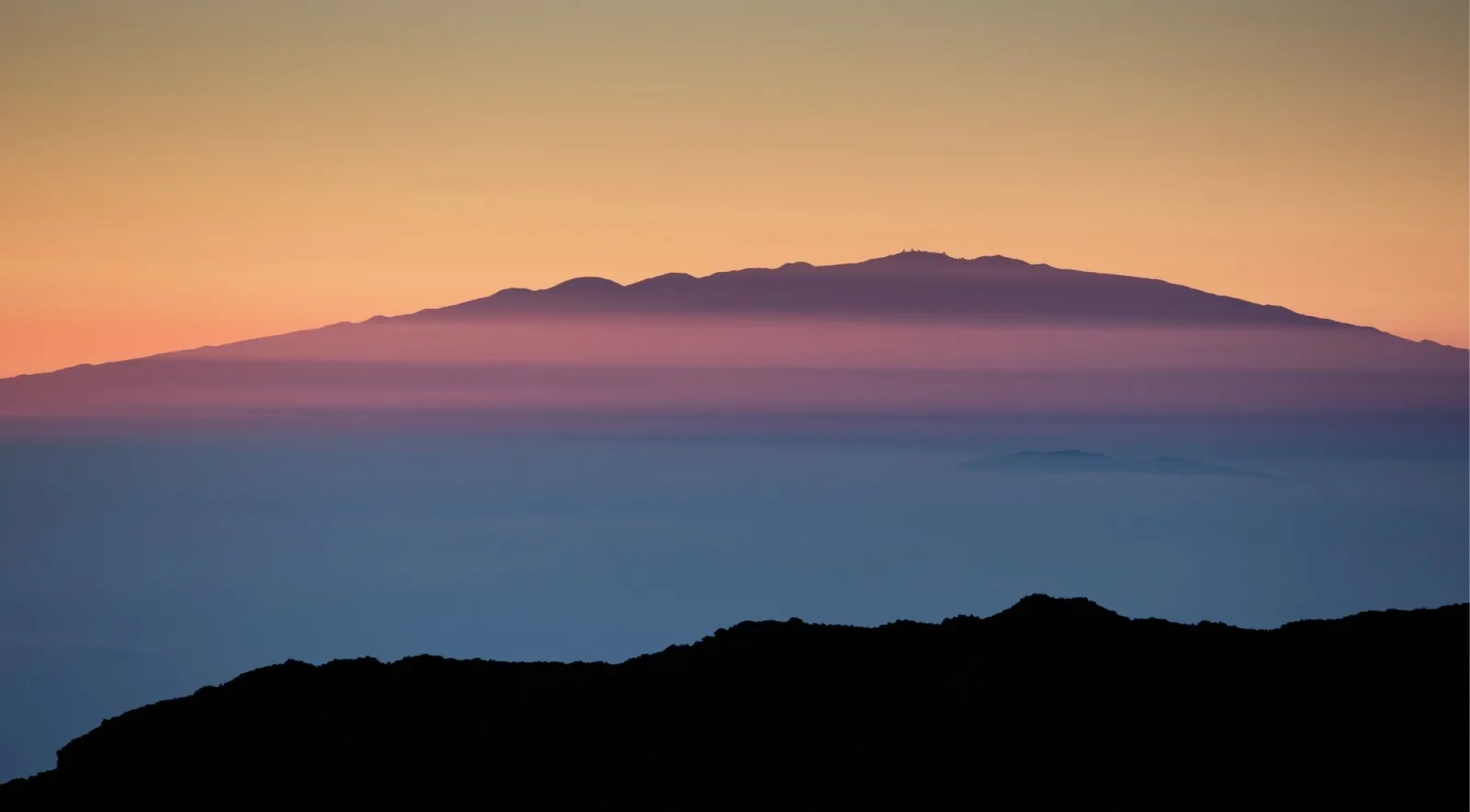 Mauna Kea silhouetted against an orange and purple sunset seen from across the Big Island, above the cloud sea