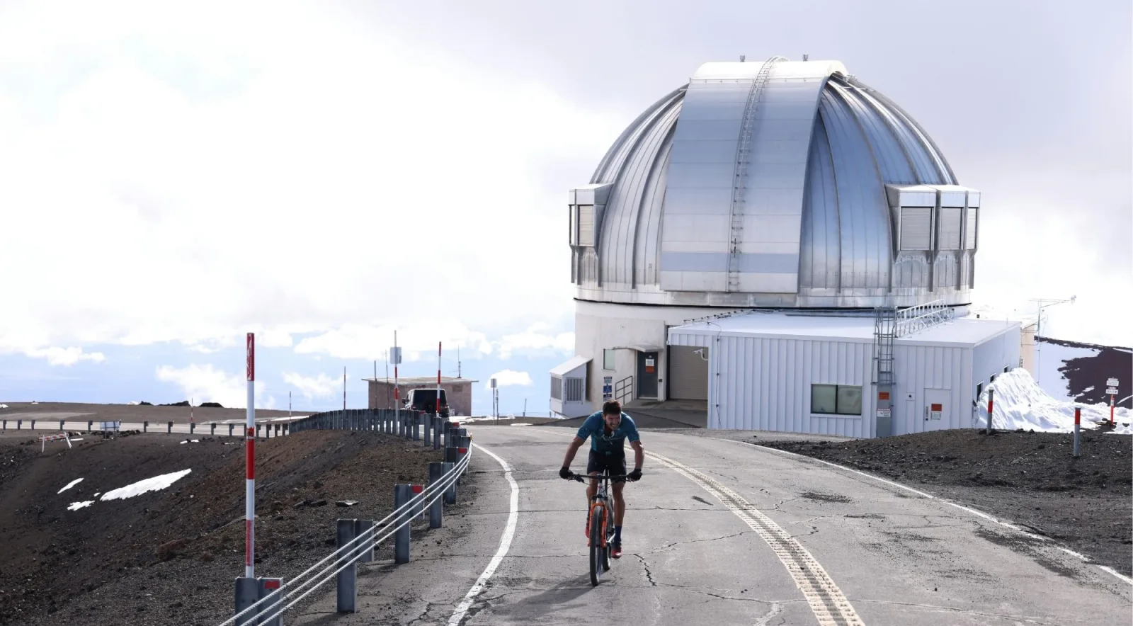 Cyclist on the Mauna Kea summit access road with a large silver observatory dome behind at 13,800 ft