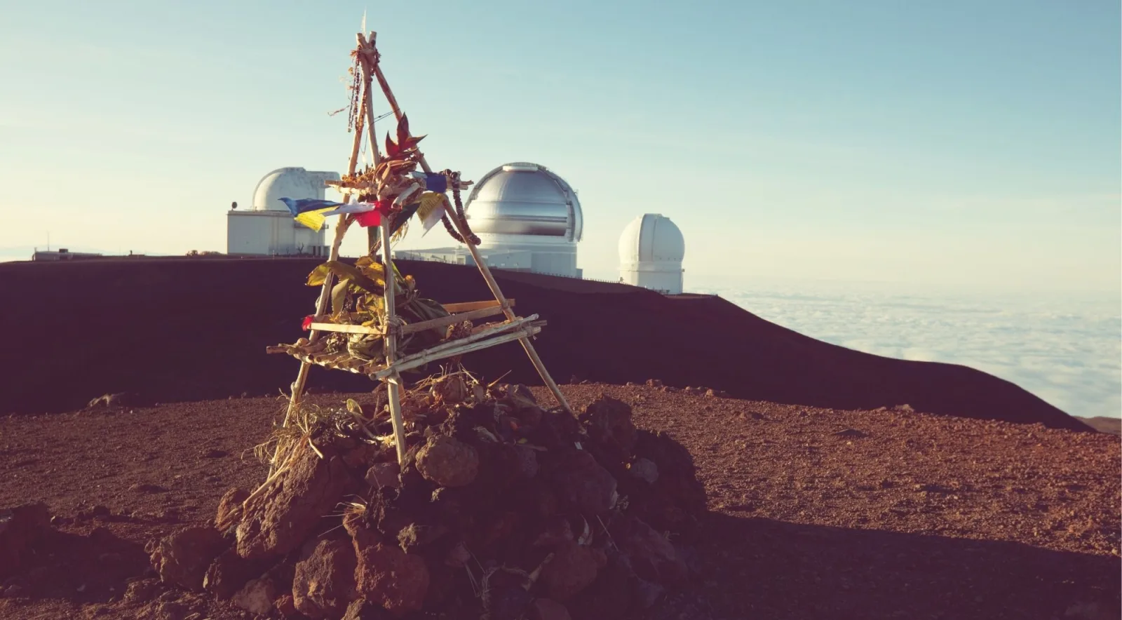 Hawaiian cultural ahu (stone altar with prayer offerings) on the Mauna Kea summit with observatories and cloud sea behind