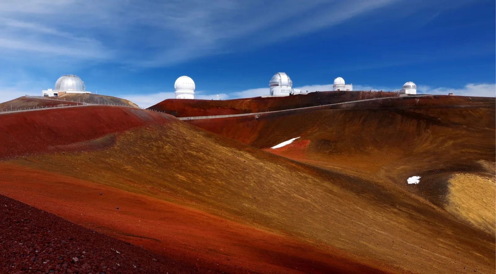 Rich red and gold volcanic cinder terrain of the Mauna Kea summit plateau with a row of observatory domes